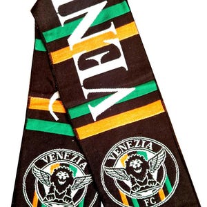 May include: A black, green, and orange knit scarf featuring the text VENEZIA in large white letters. The scarf displays two circular crests with a winged lion and the text VENEZIA FC. The ends of the scarf are finished with green and white fringe.