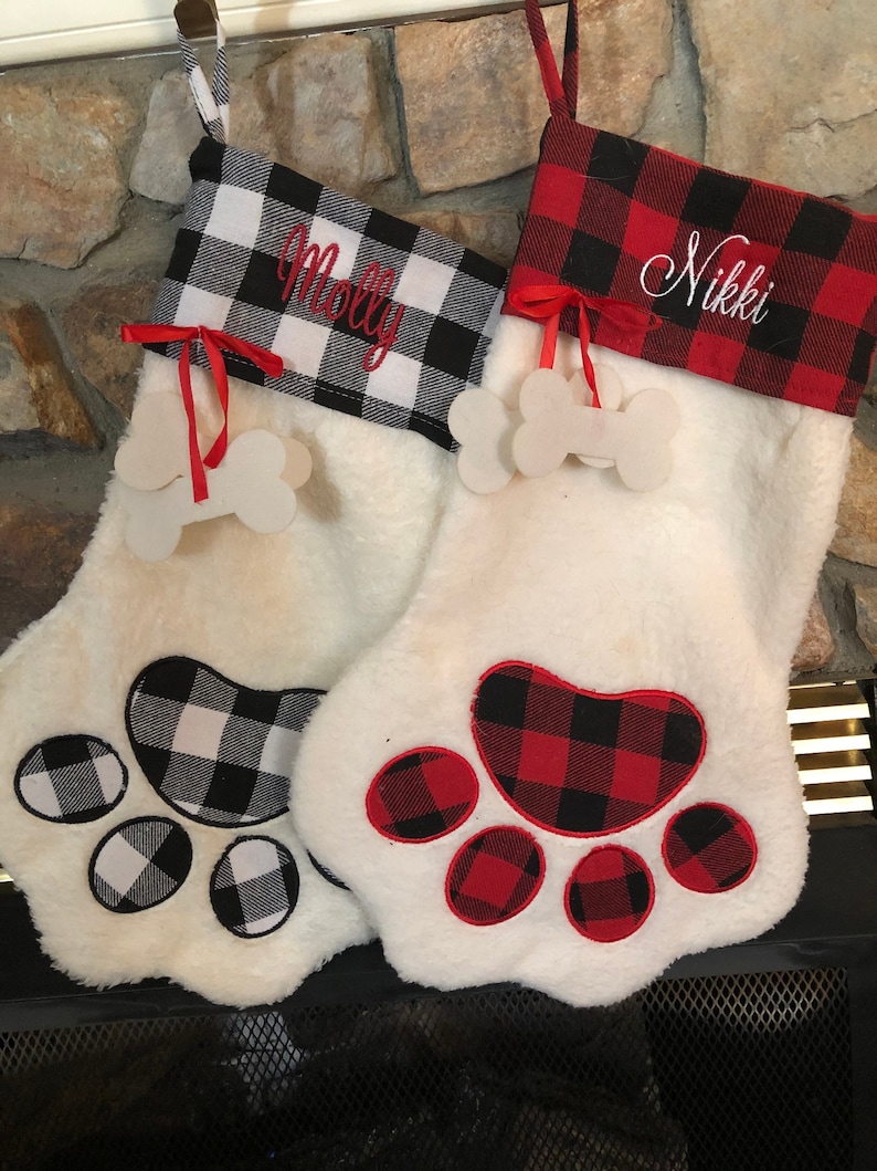 May include: Two white plush paw-shaped Christmas stockings with buffalo plaid cuffs. One cuff is black and white with the name Molly embroidered in red, the other is red and black with the name Nikki embroidered in white. Each has a bone tag.