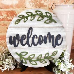 May include: A round wooden sign with a whitewashed finish and a black welcome sign. The sign is decorated with green leafy branches and is on a black stand.