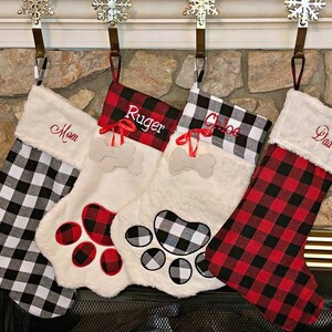 May include: Four Christmas stockings hanging from snowflake hooks. Two stockings feature black and white buffalo plaid, while two feature red and black buffalo plaid. They are personalized with the names Mom, Ruger, Chloe, and Dad.