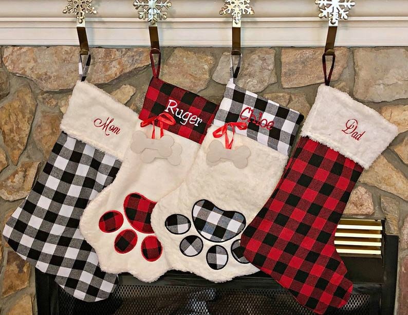 May include: Four Christmas stockings hanging from snowflake hooks. Two stockings feature black and white buffalo plaid, while two feature red and black buffalo plaid. They are personalized with the names Mom, Ruger, Chloe, and Dad.