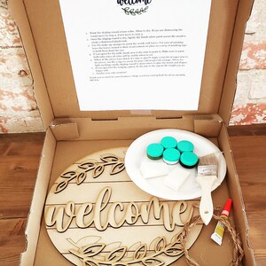 May include: A DIY farmhouse welcome sign kit with a wooden round sign, paint, brushes, and twine. The sign says "welcome" with a wreath of leaves around it.