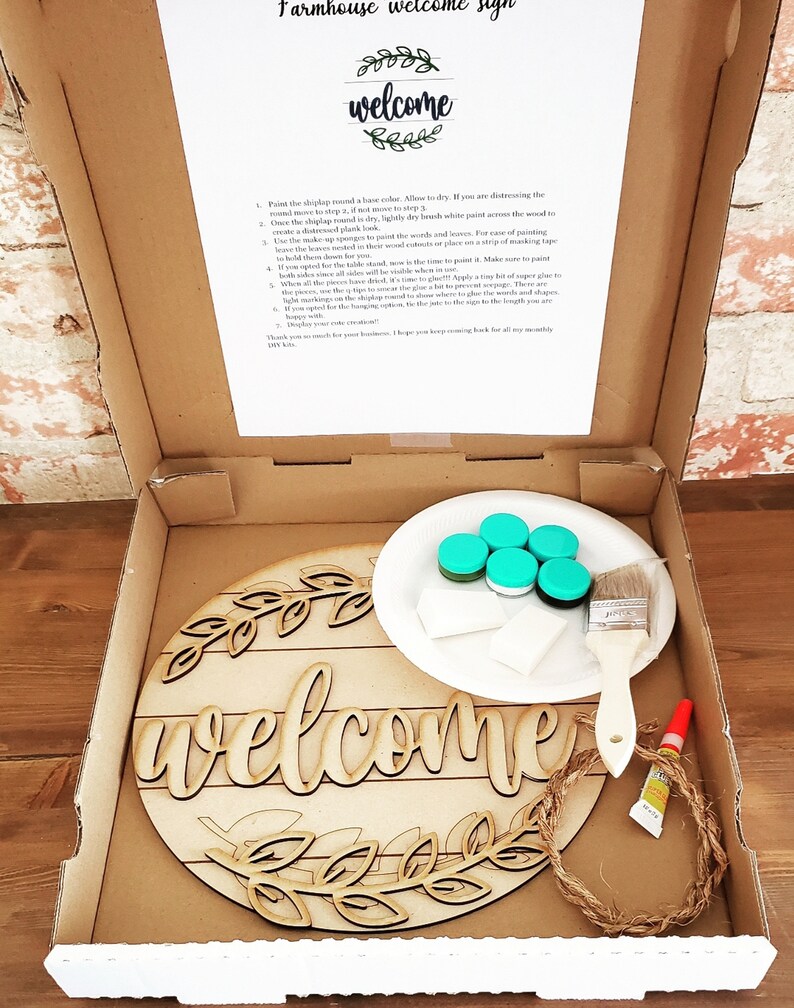May include: A DIY farmhouse welcome sign kit with a wooden round sign, paint, brushes, and twine. The sign says "welcome" with a wreath of leaves around it.