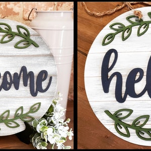 May include: Two round wooden signs with a white-washed wood grain background. One sign says "welcome" in navy blue script, and the other says "hello". Both signs have green leaf accents.