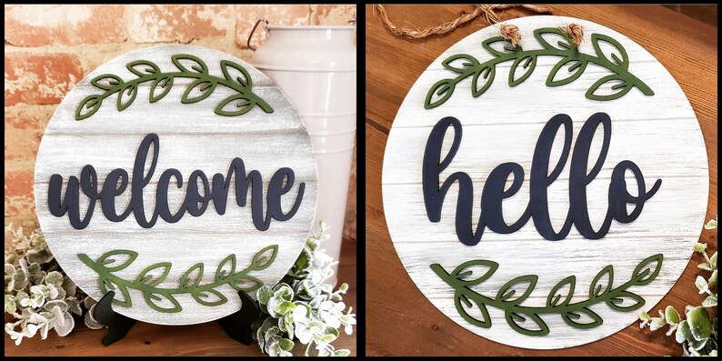 May include: Two round wooden signs with a white-washed wood grain background. One sign says "welcome" in navy blue script, and the other says "hello". Both signs have green leaf accents.