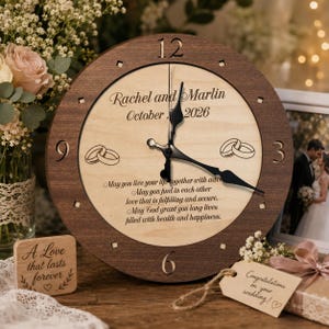 May include: A round wooden wall clock with a dark brown frame and a light wood face. The clock features black hands, engraved numbers, two interlocking ring icons, and the text: Rachel and Marlin, October 2026, and a wedding blessing.
