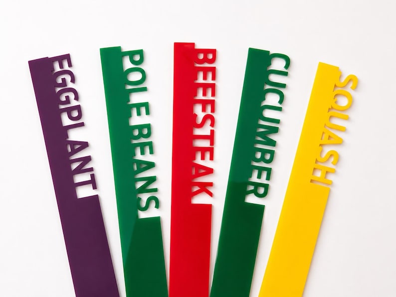 May include: Five colorful garden markers in purple, green, red, green, and yellow. Each marker has a different vegetable name cut out: Eggplant, Pole Beans, Beefsteak, Cucumber, and Squash. The markers are angled against a white background.