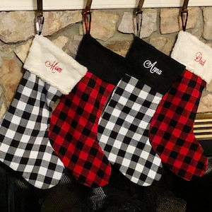 May include: Four Christmas stockings hanging in a row. Two feature a black and white buffalo check pattern with white faux fur cuffs, while two feature a red and black buffalo check pattern. The cuffs are embroidered with the names Mom, Anna, and Dad.