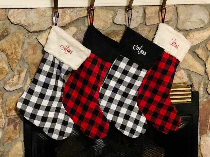 May include: Four Christmas stockings hanging in a row. Two feature a black and white buffalo check pattern with white faux fur cuffs, while two feature a red and black buffalo check pattern. The cuffs are embroidered with the names Mom, Anna, and Dad.