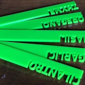 May include: Bright green herb garden markers with cut-out lettering. The markers are labeled with the names of herbs: thyme, oregano, basil, garlic, and cilantro. The markers are made of a solid, opaque material.
