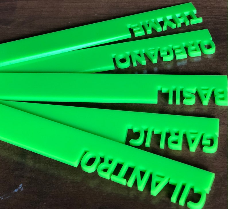 May include: Bright green herb garden markers with cut-out lettering. The markers are labeled with the names of herbs: thyme, oregano, basil, garlic, and cilantro. The markers are made of a solid, opaque material.