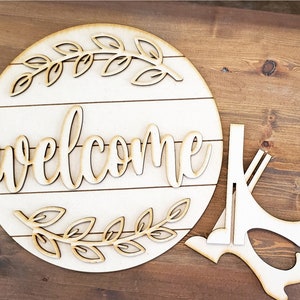 May include: A wooden welcome sign with a decorative leaf design and the word "welcome" in a cursive font. The sign is attached to a wooden stand.