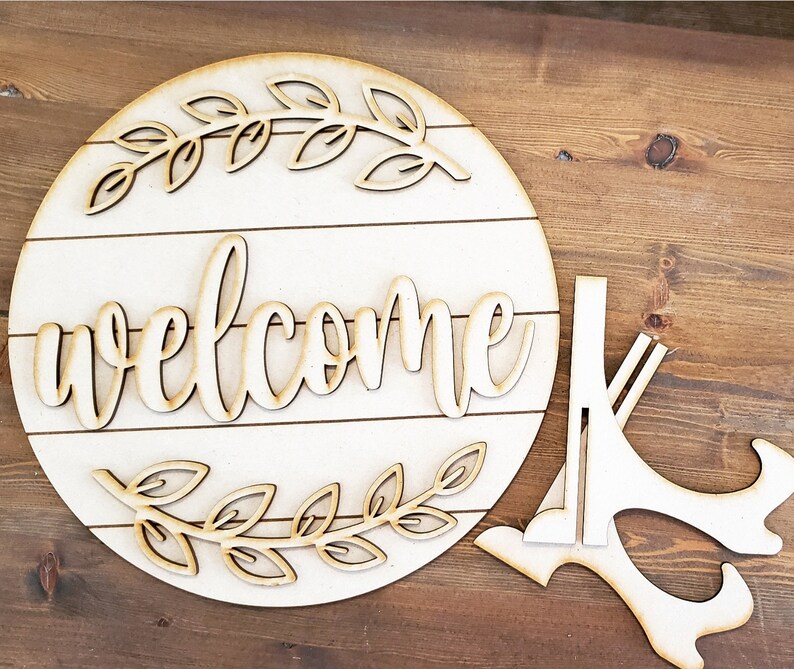 May include: A wooden welcome sign with a decorative leaf design and the word "welcome" in a cursive font. The sign is attached to a wooden stand.