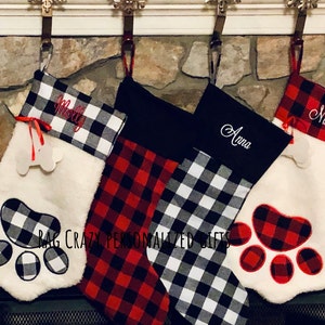 May include: Four Christmas stockings hanging in a row. Two are white faux fur with paw-shaped appliques and bone tags, one is red and black buffalo plaid, and one is black and white buffalo plaid. Names Molly, Anna, and Nikki are embroidered.