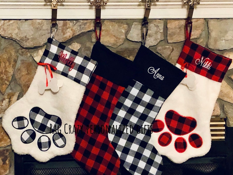 May include: Four Christmas stockings hanging in a row. Two are white faux fur with paw-shaped appliques and bone tags, one is red and black buffalo plaid, and one is black and white buffalo plaid. Names Molly, Anna, and Nikki are embroidered.