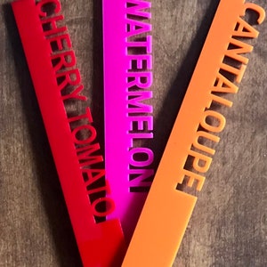 May include: Three colorful garden markers: red "Cherry Tomato", pink "Watermelon", and orange "Cantaloupe". The text is cut out of the plastic markers. These markers are designed to help identify plants in a garden.