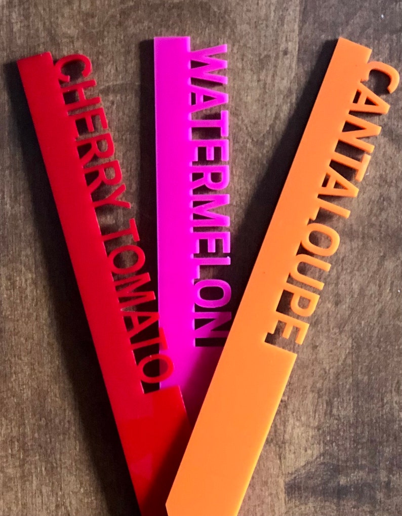 May include: Three colorful garden markers: red "Cherry Tomato", pink "Watermelon", and orange "Cantaloupe". The text is cut out of the plastic markers. These markers are designed to help identify plants in a garden.
