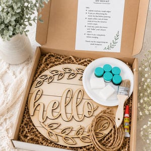 May include: A craft kit for a 'hello' sign in a cardboard box. The kit contains a wooden sign with the word 'hello', paint, a brush, glue, and rope. The sign features a leaf design. Instructions are also included.