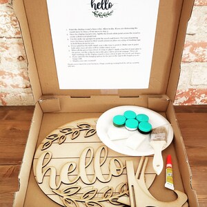 May include: A DIY farmhouse sign kit with a wooden round sign, paint, brushes, and glue. The sign says "hello" with a wreath of leaves around it.