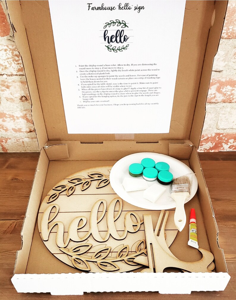 May include: A DIY farmhouse sign kit with a wooden round sign, paint, brushes, and glue. The sign says "hello" with a wreath of leaves around it.