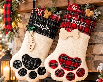 May include: Two paw-shaped Christmas stockings made of white faux sherpa fabric. The left stocking features a black and green plaid cuff with the name Murphy and a wooden fish bone tag. The right stocking has a red and black plaid cuff with the name Buzz and a wooden bone tag.