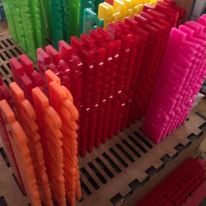 May include: A collection of colorful, interlocking plastic pieces in shades of orange, red, yellow, green, and pink. The pieces are arranged in rows within a wooden tray, suggesting a building or design project. The pieces have a textured surface.