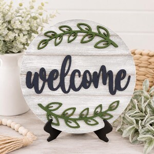 May include: A circular wooden sign featuring the word "welcome" in navy script. The sign has a white-washed wood grain effect and is decorated with green leaf accents. It is displayed on a black easel.
