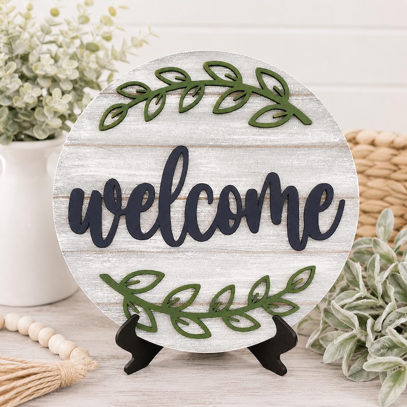 May include: A circular wooden sign featuring the word "welcome" in navy script. The sign has a white-washed wood grain effect and is decorated with green leaf accents. It is displayed on a black easel.