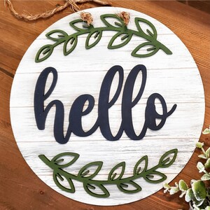 May include: A round wooden sign with a whitewashed finish and a distressed look. The sign features a black cursive word "hello" surrounded by two green leafy wreaths.