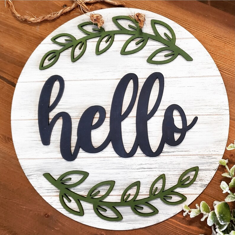 May include: A round wooden sign with a whitewashed finish and a distressed look. The sign features a black cursive word "hello" surrounded by two green leafy wreaths.