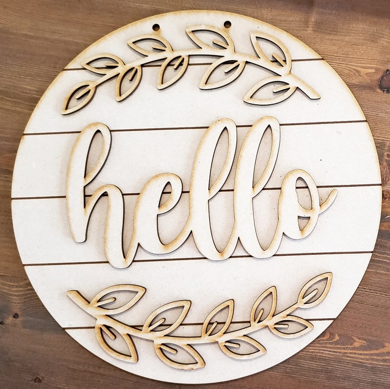 May include: A round wooden sign with a white background and the word "hello" written in a cursive font. The sign is decorated with two wreaths of leaves.