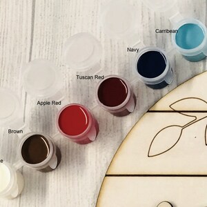 May include: Close-up of paint pots in various colours, including brown, apple red, and navy. The paint pots are labelled with their respective colours. A wooden craft piece with leaf cutouts is partially visible.