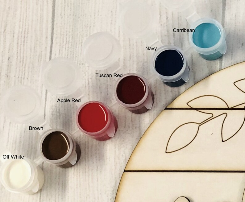 May include: Close-up of paint pots in various colours, including brown, apple red, and navy. The paint pots are labelled with their respective colours. A wooden craft piece with leaf cutouts is partially visible.