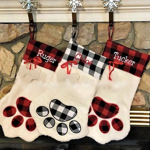 May include: Three white plush pet stockings shaped like paws. Two stockings feature red and black buffalo plaid cuffs with the names Ruger and Tucker, while the center one has a black and white plaid cuff with the name Chloe.