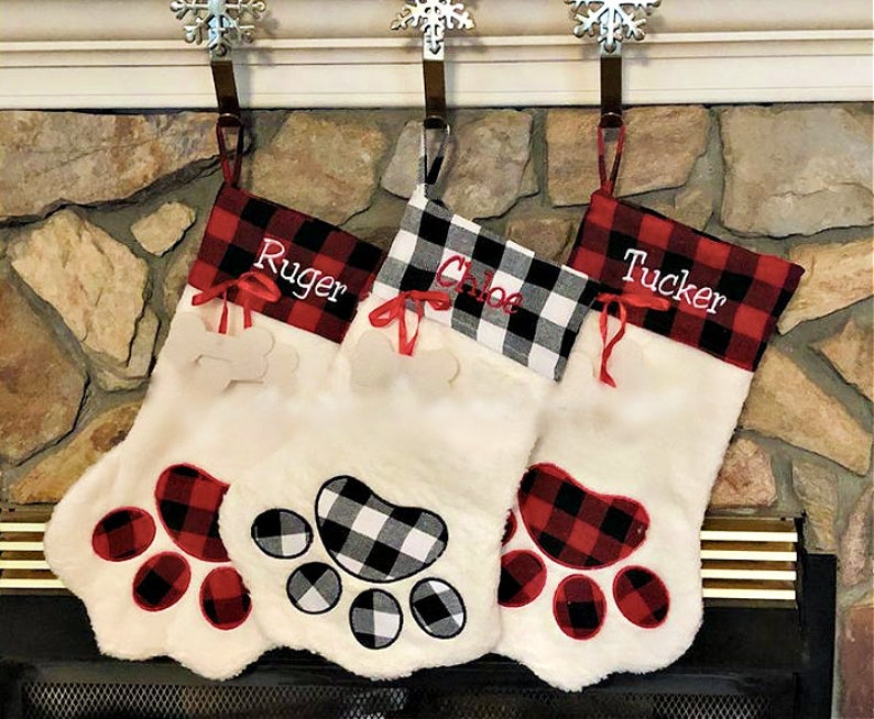 May include: Three white plush pet stockings shaped like paws. Two stockings feature red and black buffalo plaid cuffs with the names Ruger and Tucker, while the center one has a black and white plaid cuff with the name Chloe.