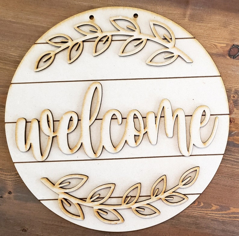May include: Circular wooden welcome sign with a white base and natural wood detailing. The word "welcome" is in a cursive font, with leaf accents. The sign is designed to be hung.