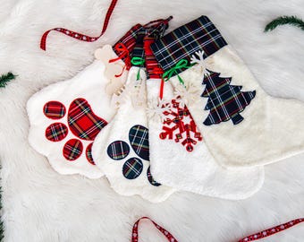 May include: Four white Christmas stockings with plaid accents and decorative embellishments. One stocking features a paw print design, while another has a Christmas tree. Red and green ribbons and faux greenery add to the festive theme.