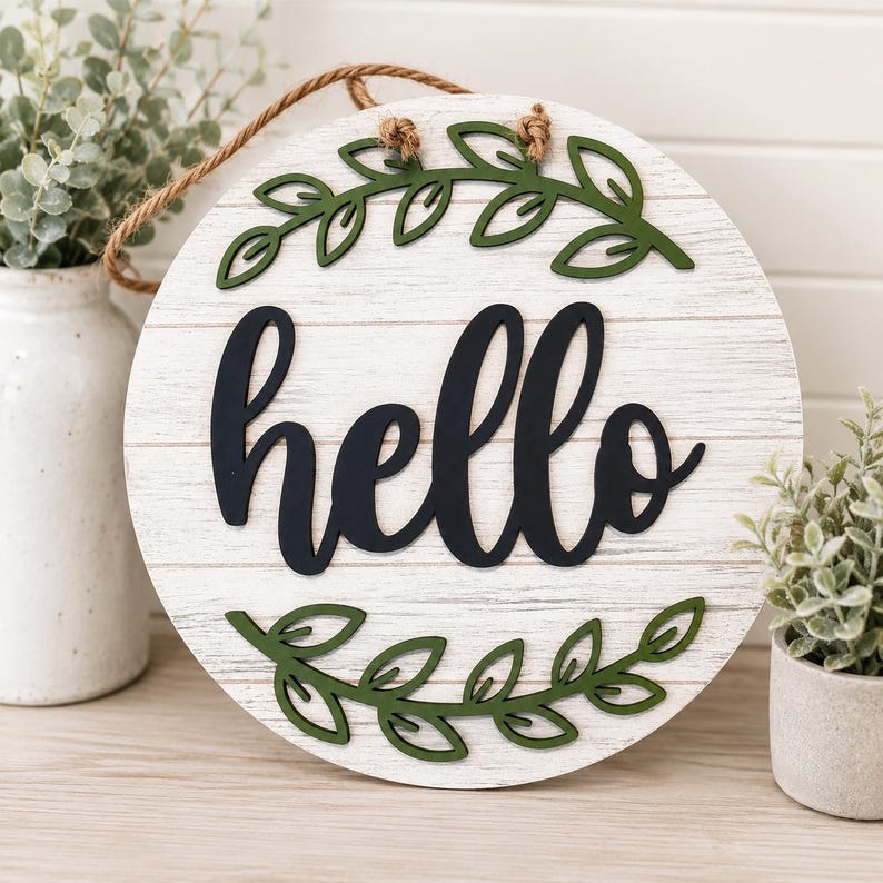 May include: A circular wooden sign with a white-painted, distressed finish, featuring the word "hello" in black script. Green leaf detailing adorns the top and bottom. A natural rope is attached for hanging.