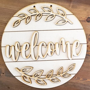 May include: A round wooden sign with a white background and the word "welcome" in a cursive font. The sign is decorated with two wreaths of leaves.