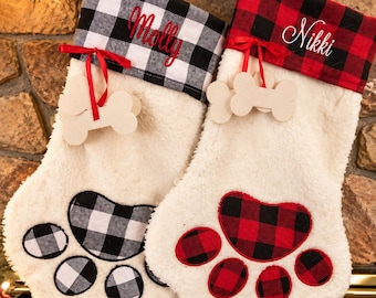 May include: Two paw-shaped Christmas stockings made of cream-colored faux fur. One features a black and white buffalo check cuff with the name Molly, the other a red and black buffalo check cuff with the name Nikki. Both have bone-shaped tags.