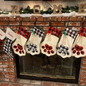 May include: Seven Christmas stockings hanging in a row, featuring a mix of red and black or white and black buffalo plaid patterns. Five stockings are shaped like animal paws with plush white fabric and personalized names embroidered.