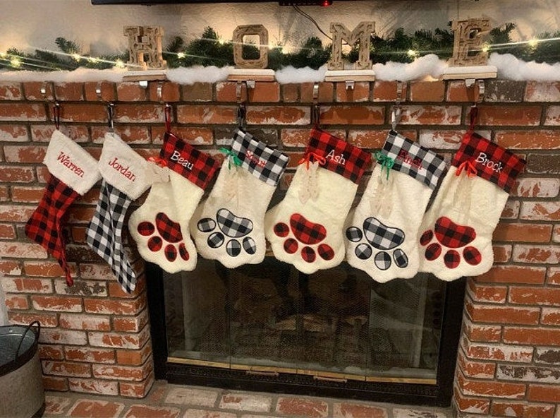 May include: Seven Christmas stockings hanging in a row, featuring a mix of red and black or white and black buffalo plaid patterns. Five stockings are shaped like animal paws with plush white fabric and personalized names embroidered.