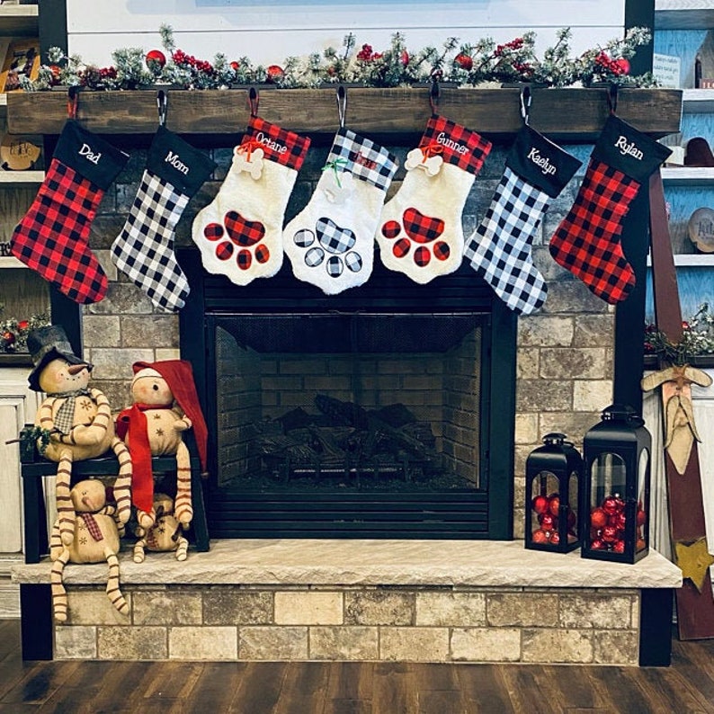 May include: Seven Christmas stockings hang from a wooden mantel. Two are red and black buffalo plaid, two are white and black buffalo plaid, and three are white faux fur paw-shaped stockings with plaid accents and names embroidered on the cuffs.