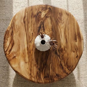 Peut inclure: Une table basse ronde en bois avec un bord naturel, présentant des motifs de grain de bois brun et beige. Un petit vase en céramique blanche avec des fleurs séchées est posé sur la table. Le diamètre de la table est d'environ 122 cm.