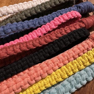 May include: Close-up of several macrame wrist straps in various colors, including white, olive green, blue, pink, red, black, coral, yellow, and light blue. Each strap is made of woven cotton cord and has a metal clasp.