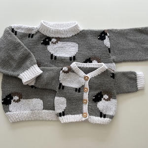 Könnte beinhalten: Ein grauer Strickpullover und eine passende graue Strickjacke, beide mit einem wiederkehrenden Muster aus weißen Schafen mit schwarzen Gesichtern und braunen Hörnern. Die Kleidungsstücke haben weiße Rippbündchen an Kragen, Ärmeln und Saum sowie vier Holzknöpfe.