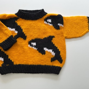Puede incluir: Un suéter de punto de manga larga en color amarillo mostaza vibrante con un patrón de intarsia repetitivo de orcas negras y blancas. La prenda tiene un cuello redondo negro acanalado, puños negros acanalados y un dobladillo negro acanalado.
