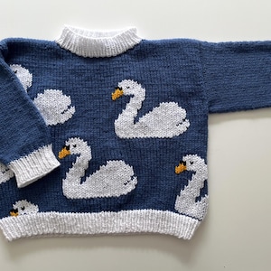 May include: A long-sleeved knitted jumper in deep blue featuring a repeating pattern of white swans with orange beaks. The garment has a ribbed white crew neckline, white ribbed sleeve cuffs, and a wide white ribbed hem at the bottom edge.