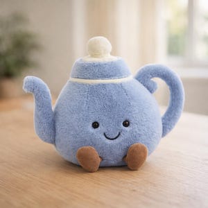 Puede incluir: Una tetera de peluche azul claro con una cara sonriente, ojos negros y pequeños pies marrones. La tetera tiene un borde blanco y un botón blanco en la tapa. El asa y el pico son curvos. El juguete está sobre una superficie de madera.