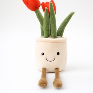 以下が含まれることがあります： 笑顔の鉢植えのぬいぐるみ。茶色の足があり、赤いチューリップが3つ、緑の茎と葉が付いています。オフホワイトの鉢は柔らかく、質感のある生地で作られています。魅力的で気まぐれなデザインです。